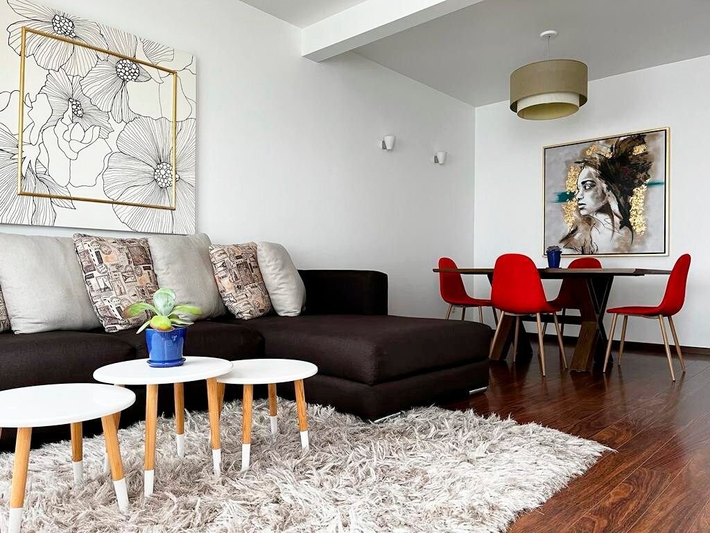 A modern living area features a dark sectional sofa adorned with decorative pillows. A soft rug lies on the polished floor, complementing a small table with a potted plant. A dining table with red chairs is visible in the background, highlighted by a large artwork on the wall.
