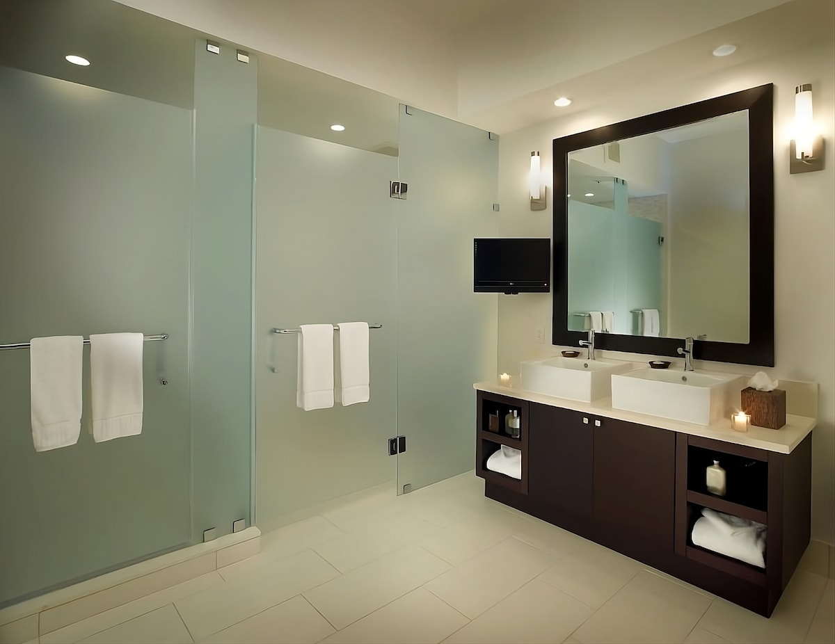 A modern bathroom features dual sinks integrated into a sleek countertop. Clear glass shower partitions define the space, complemented by soft lighting and neatly arranged towels. The layout promotes an open ambiance, enhancing the overall functionality and aesthetics of the area.