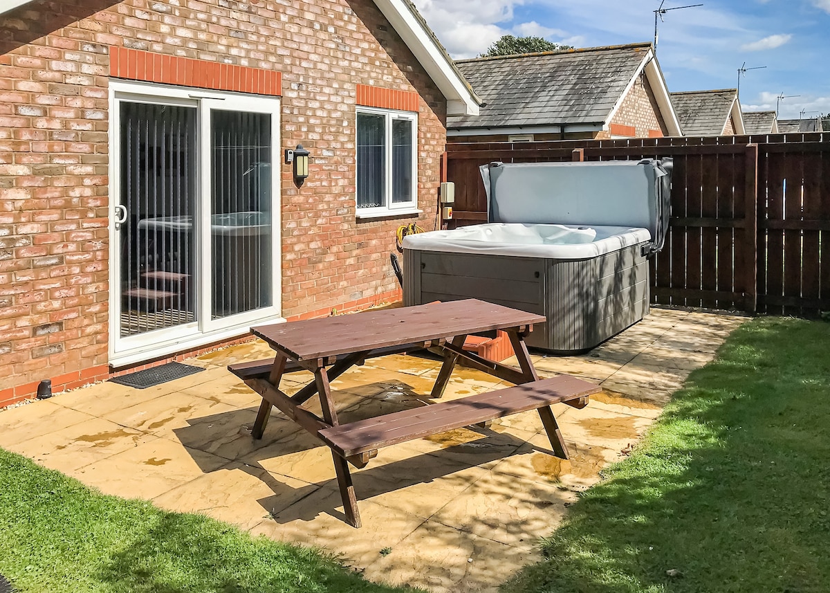 A private outdoor space is featured, with a wooden picnic table positioned on a stone patio. A hot tub is seen next to a wooden fence, while grassed areas provide a natural setting. Large sliding doors lead into the cottage.