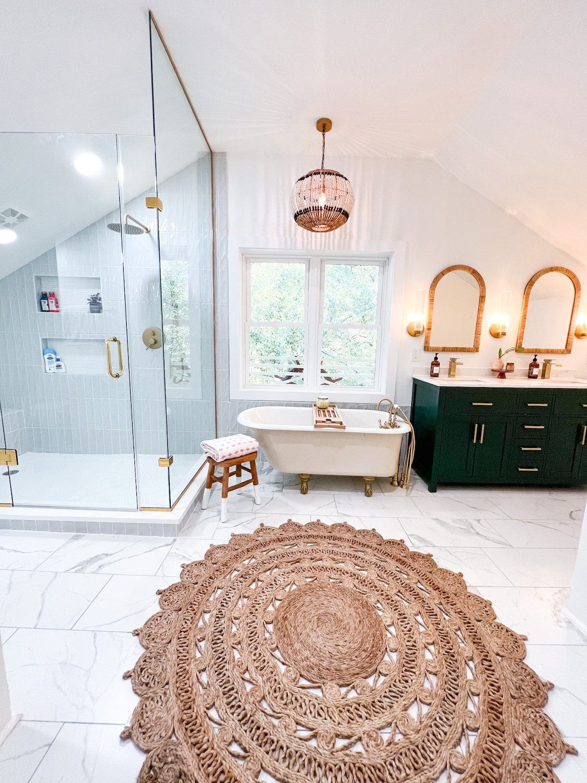 A spacious bathroom features a glass-enclosed shower with a rain showerhead and a standalone bathtub. Dual mirrors are mounted above a dark green vanity. Natural light enters through a large window, illuminating the stylish decor, including a round, woven rug on the marble floor.