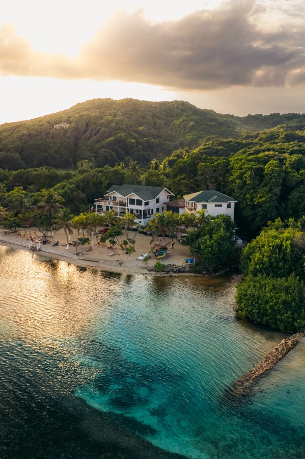 An aerial view captures a resort nestled along a sandy beach, surrounded by lush greenery. The coastline showcases calm waters reflecting the colors of the setting sun, while the resort's structures blend harmoniously with the natural landscape.