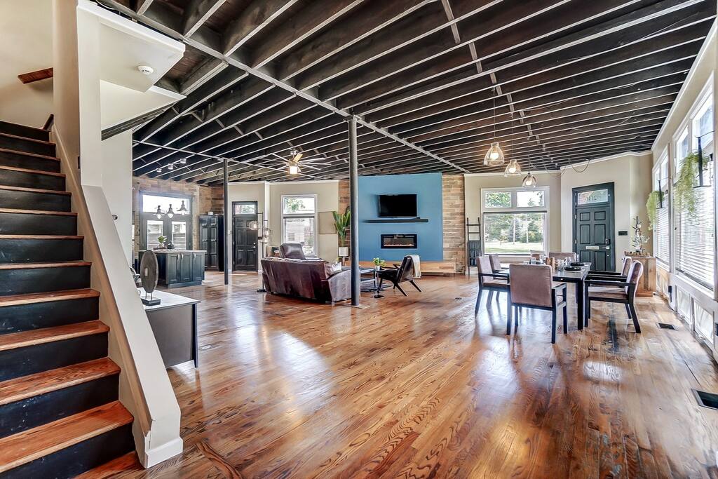 An open living space features soaring ceilings with exposed beams and large windows allowing natural light. The floor is finished in warm wood, and modern furniture is arranged for comfort. A staircase leads to the upper levels, while a dining area is positioned near the windows.