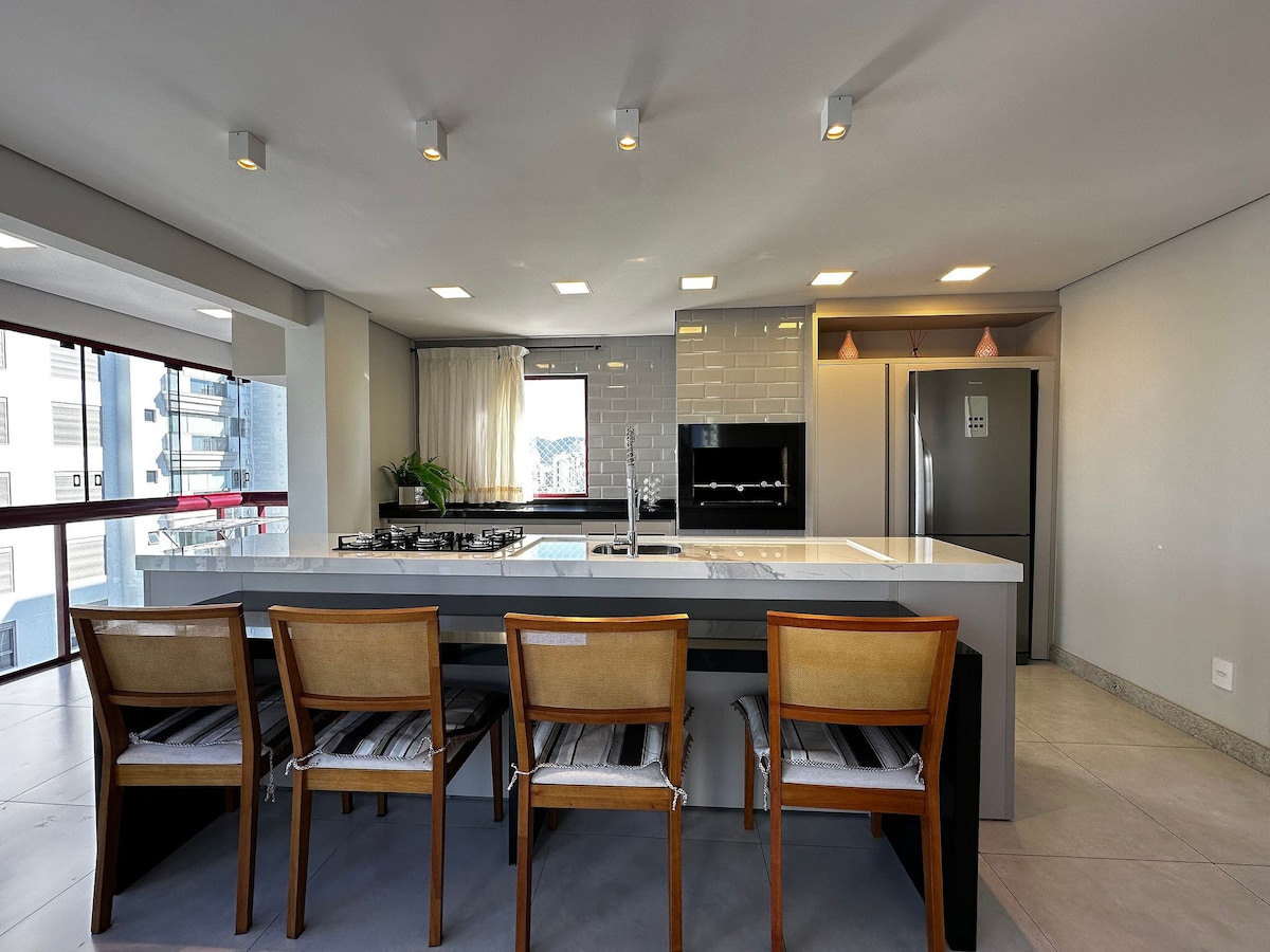 The modern kitchen features a large island with ample countertop space, complemented by a minimalist gas stove. Bar stools offer seating along one side, while large windows provide natural light and views. A built-in grill is also visible, enhancing the functionality of the space.