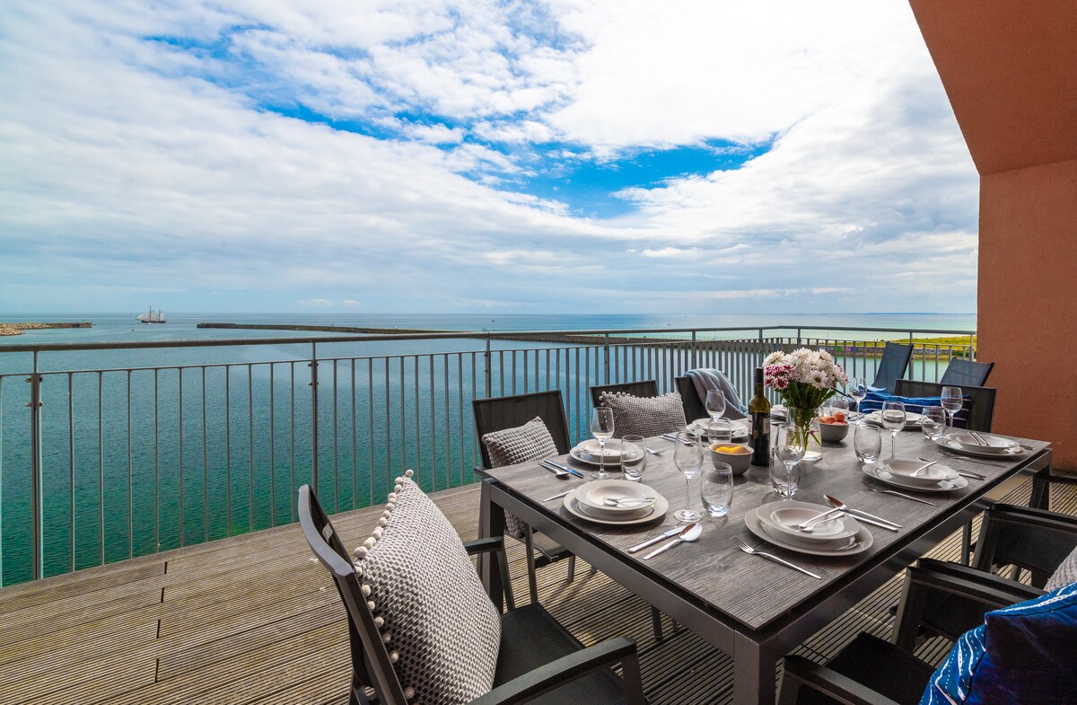 A spacious terrace features a dining table set for a meal with a view. The table is adorned with plates, glasses, and a vase of flowers. Beyond, the calm sea extends to the horizon under a partly cloudy sky, inviting relaxation.