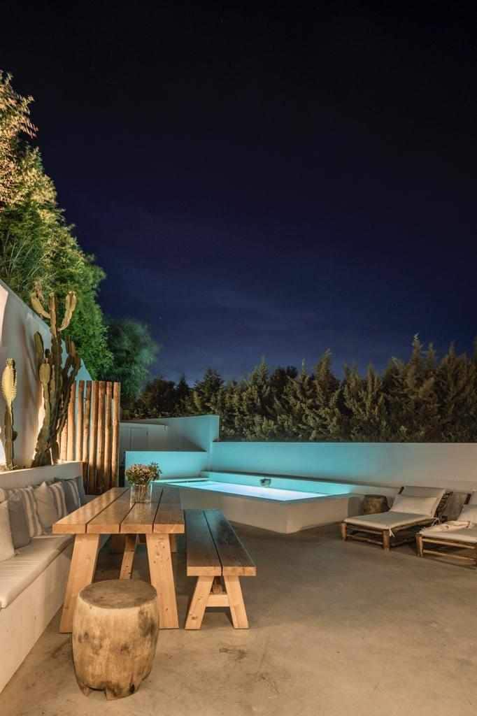 An outdoor patio is illuminated at night, featuring a swimming pool and a long wooden dining table surrounded by comfortable seating. The area is complemented by lush greenery and a serene atmosphere, inviting relaxation under the evening sky.