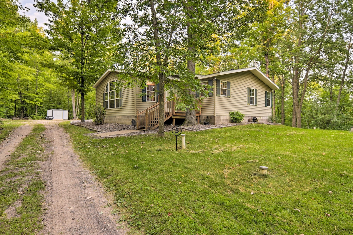 A spacious cabin is nestled among lush greenery, surrounded by trees. A dirt path leads up to the entrance, where steps are visible. The cabin features numerous windows that provide natural light and offer views of the forested surroundings.