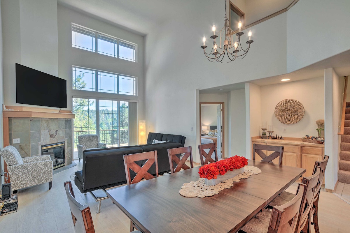 A spacious dining area features a large wooden table surrounded by eight chairs. Natural light streams in from tall windows, illuminating the room. A neutral-toned couch is visible in the background, along with a modern chandelier. A stone fireplace adds warmth to the space.