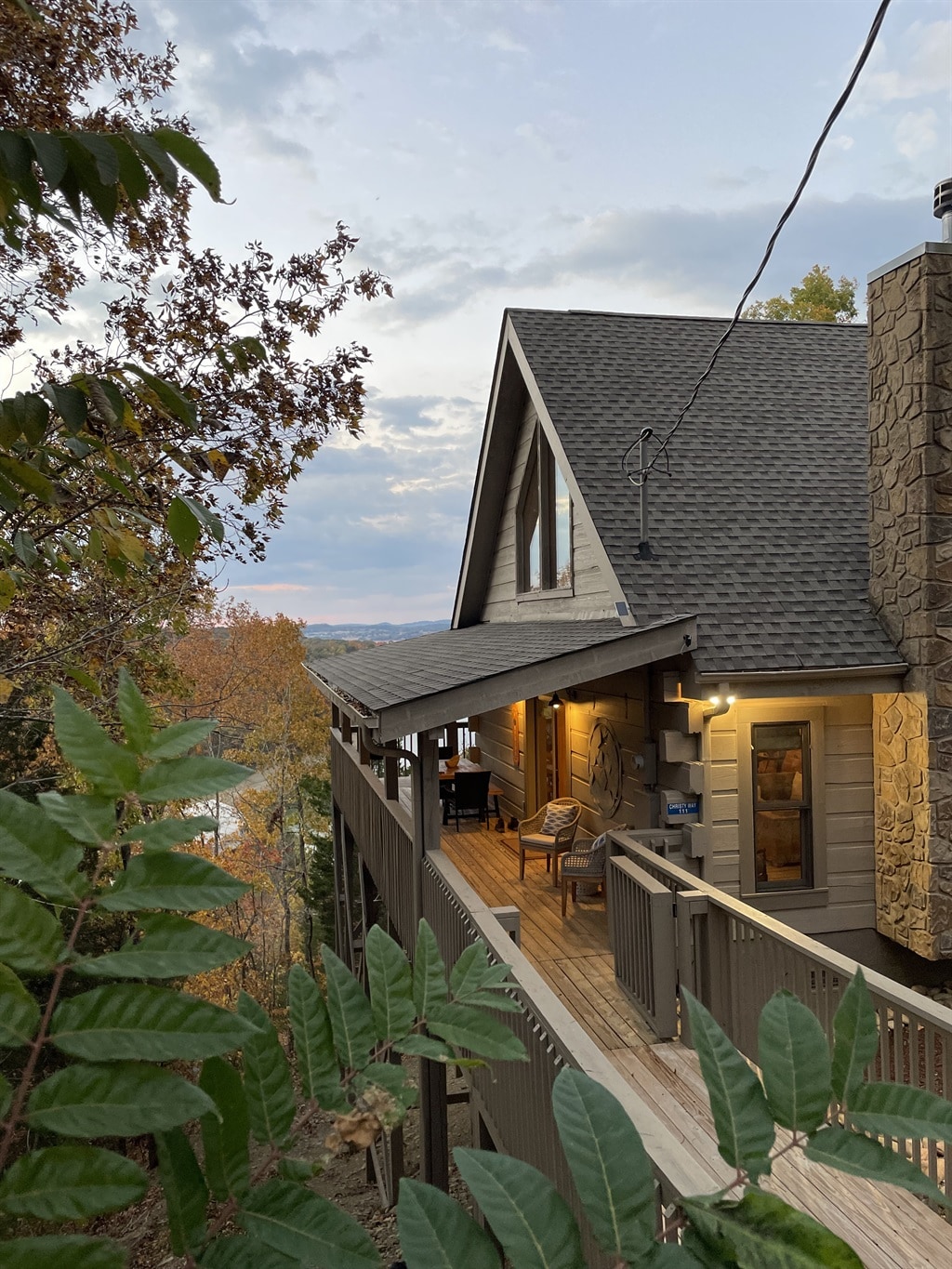The exterior of the cabin is framed by leafy greenery, showcasing a spacious deck leading to the entrance. Natural stone accents enhance the cabin's facade, while the sloped roof adds architectural interest. A serene lakeside view can be glimpsed through the trees.