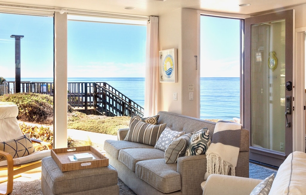 An inviting living area is highlighted by large windows that offer unobstructed ocean views. A comfortable sofa adorned with decorative pillows faces the serene coastline. Natural light fills the space, creating a bright atmosphere. The room connects seamlessly to the outdoor access, emphasizing the beach proximity.