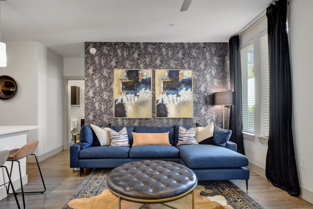 A well-furnished living area features a deep blue sectional sofa adorned with colorful cushions. Two abstract paintings hang above the couch, complemented by a round coffee table on a patterned area rug. Natural light enters through window blinds, enhancing the modern decor.