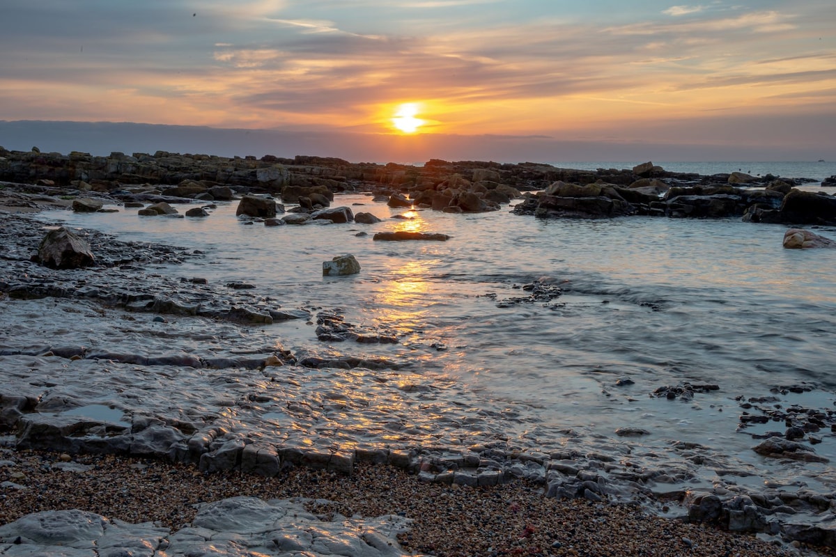 A tranquil seascape is presented at sunset, with the sun dipping below the horizon. Reflections shimmer on the calm water, framed by rocky shorelines and smooth pebbles. The sky showcases a soft gradient of warm hues, enhancing the serene coastal atmosphere.