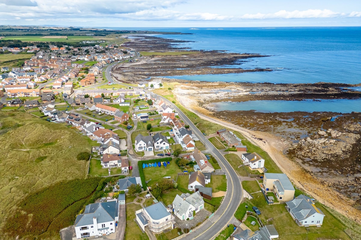 An aerial view captures the charming coastal village of Beadnell, showcasing the local homes and the serene ocean. The shoreline is composed of rocky outcrops, while sandy areas are visible. The surrounding landscape features green fields, with a clear view of the sea stretching into the horizon.