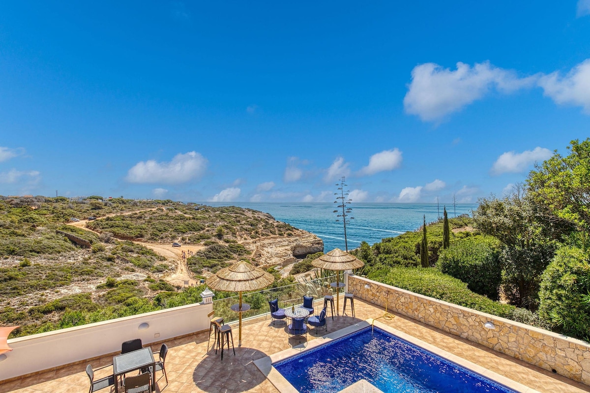 An outdoor space showcases a private swimming pool bordered by sun loungers and shaded areas. The serene ocean view complements the surrounding natural landscape, featuring cliffs and greenery under a clear blue sky. The tranquil environment is ideal for relaxation and enjoying the vistas.