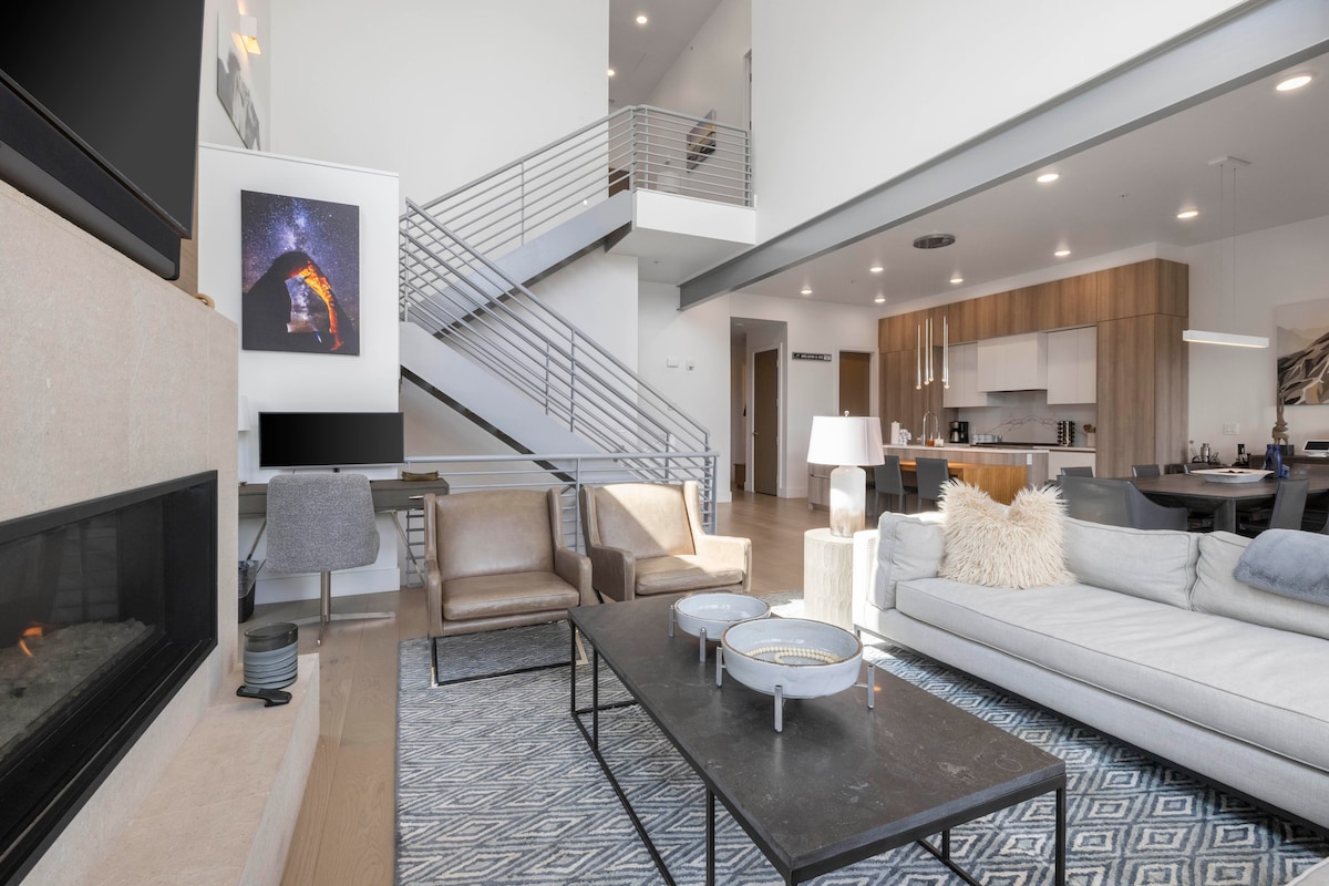 A spacious living area is designed with high ceilings and floor-to-ceiling windows allowing ample natural light. A modern fireplace is flanked by a sectional sofa and armchairs arranged around a flat coffee table. A sleek kitchen is visible in the background, accessible via an open layout.