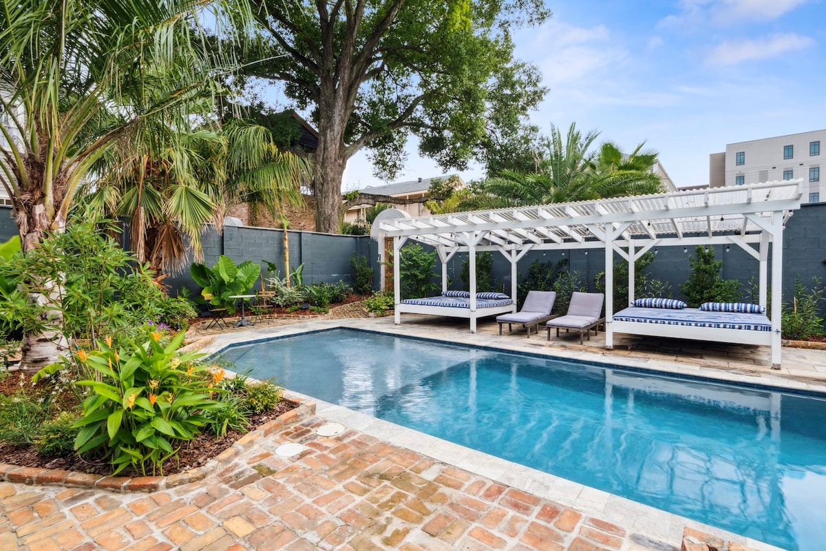 A private outdoor space includes a sparkling heated pool surrounded by lush greenery. A wooden pergola offers shaded seating with striped daybeds, while brick pathways lead to tropical plants. The area is designed for relaxation and outdoor dining in a serene setting.