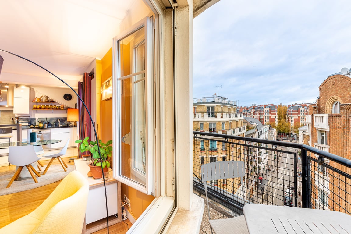 A balcony view reveals a vibrant urban landscape, with architectural features contrasting against the sky. The inviting interior is visible, showcasing a dining area with modern seating and a potted plant. The large window allows natural light to brighten the space.