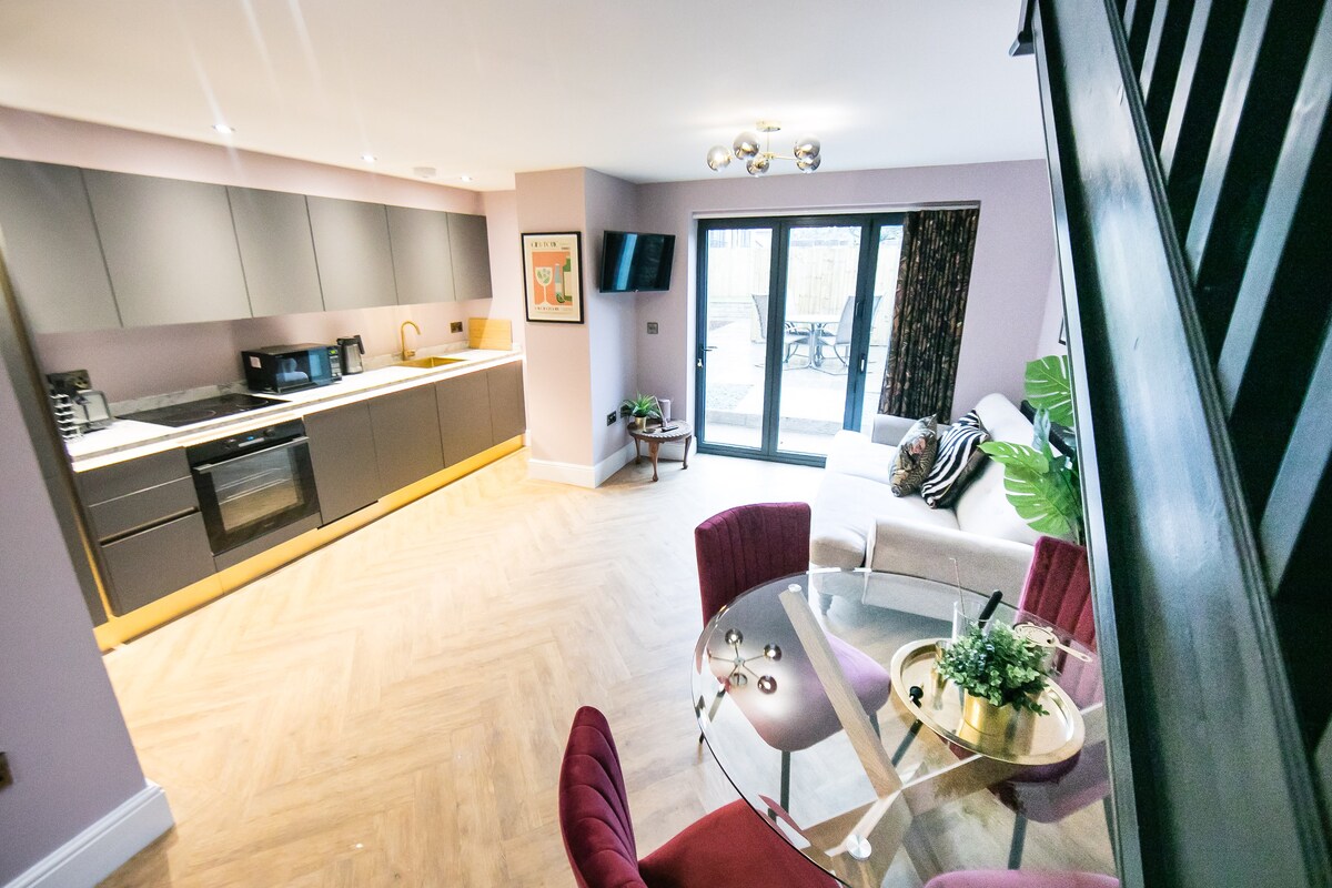 A modern kitchen and living area feature sleek cabinetry and appliances. A glass dining table is surrounded by red upholstered chairs. A comfortable sofa is placed nearby, with patio doors leading to an outdoor area, allowing natural light to fill the space.