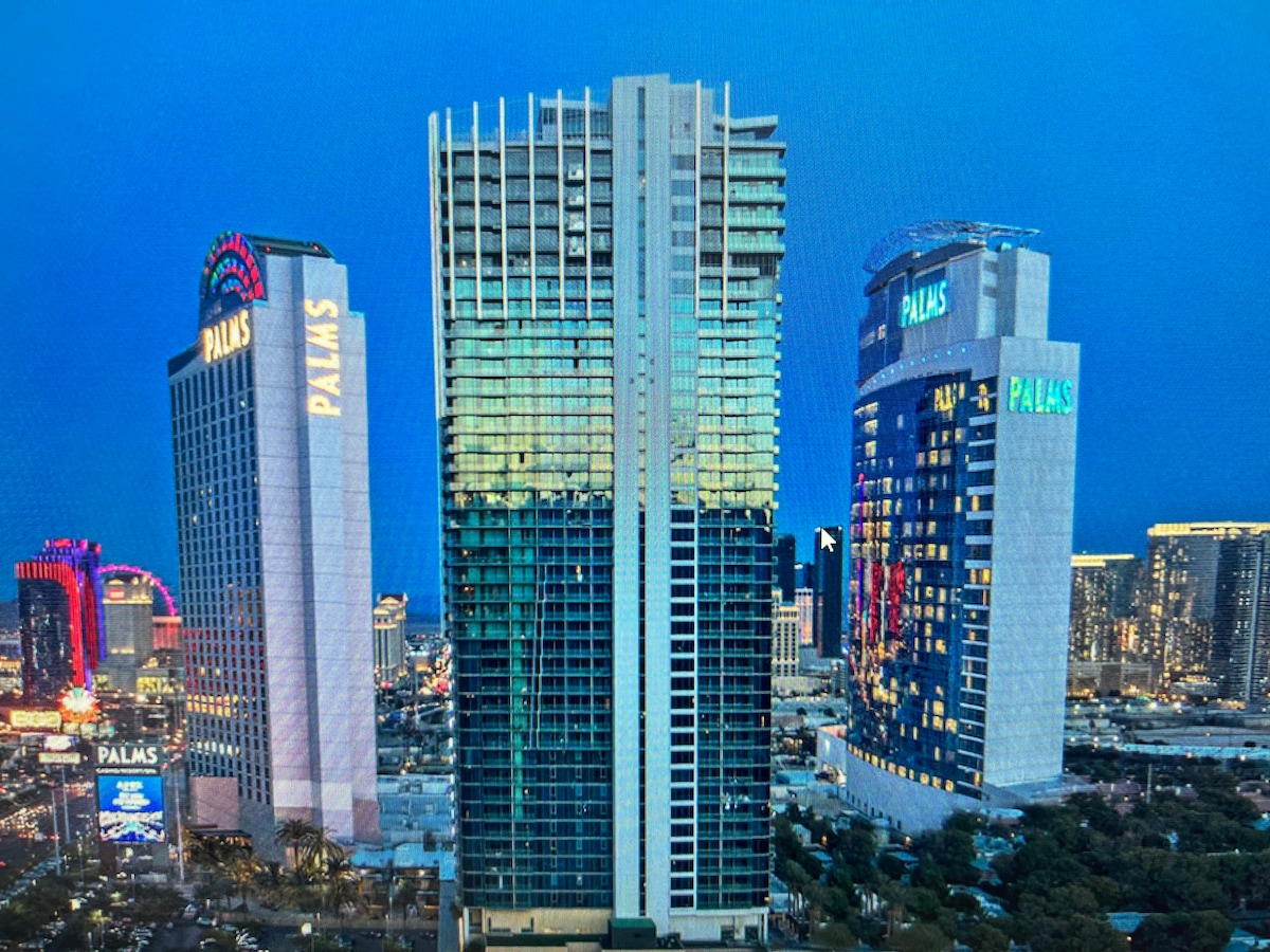The Palms Place building is showcased in an evening setting, reflecting lights from the surrounding Las Vegas Strip. The structure is designed with a sleek modern façade, featuring large glass windows and illuminated signage, blending into the vibrant atmosphere of the city.