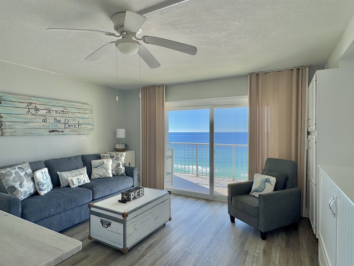 A welcoming living space features a comfortable navy sofa adorned with decorative pillows. A coffee table is positioned centrally in front of the sofa. Natural light floods the area through large glass doors, which offer a stunning view of the ocean and balcony.