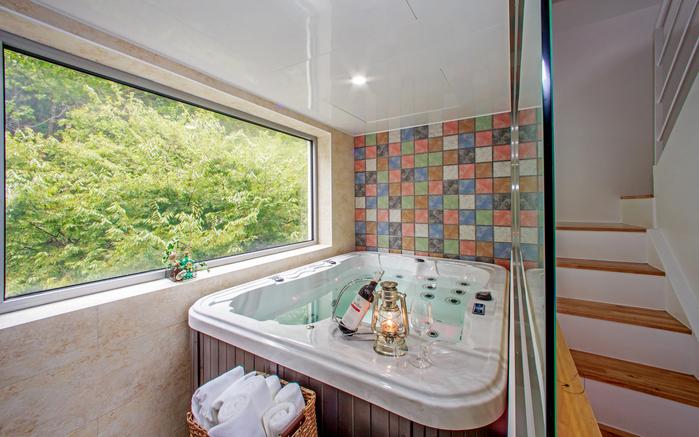 A well-appointed hot tub is framed by a large window, offering views of lush greenery. The tiled wall features a colorful mosaic design. A decorative lantern and glasses are placed nearby, adding to the serene atmosphere.