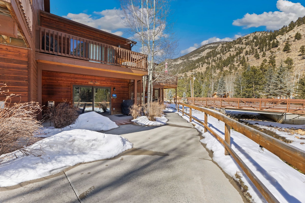 An exterior view of the building showcases a private deck with patio furniture, surrounded by scenic winter landscapes. Snow blankets the ground, and nearby mountains create a stunning backdrop, highlighting a peaceful environment.