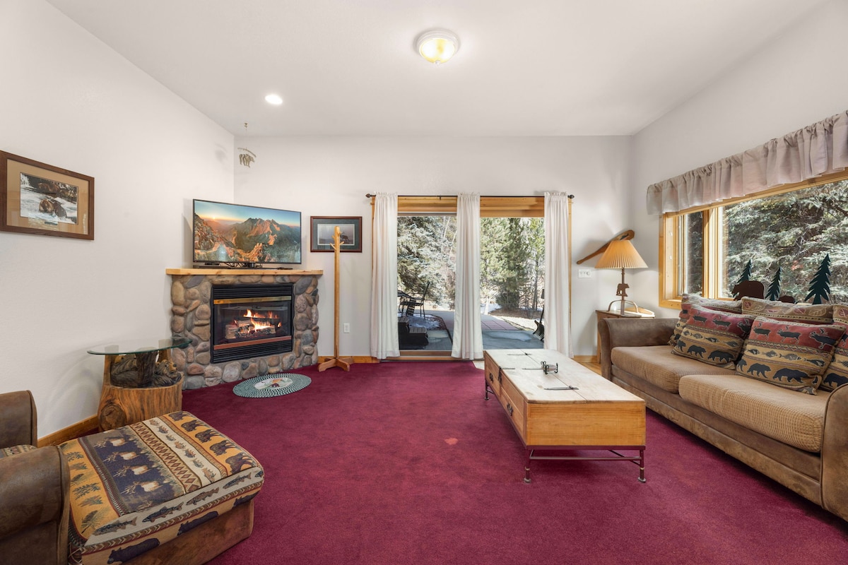 A living room area features a cozy gas fireplace surrounded by stone, with a wooden coffee table positioned in front. A large window offers views of a forested exterior, and light enters through sliding glass doors that lead to the private patio.