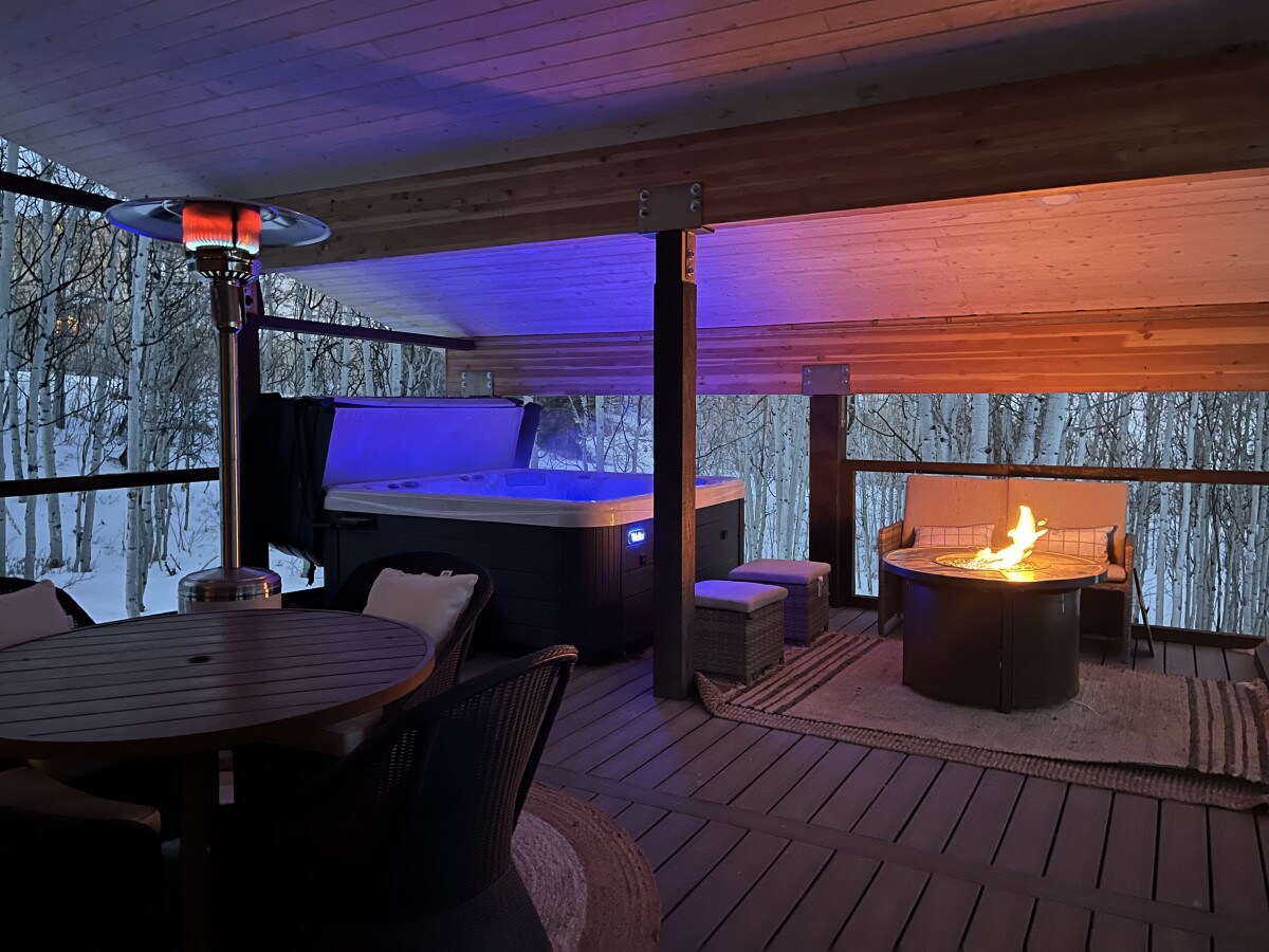 An inviting semi-enclosed balcony is featured, showcasing a hot tub and a fire pit with a warm flame. Surrounding seating areas provide a comfortable setting, while the wooden ceiling and wide wooden flooring create a cozy ambiance. Snow-covered trees are visible in the background.