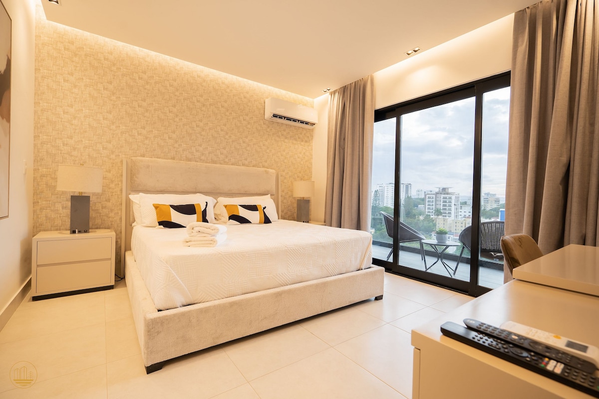 A modern bedroom features a spacious bed adorned with crisp white linens. Large windows provide natural light and a view of the surrounding city. Neutral tones complement the minimalist décor, while two bedside lamps add functionality. A small seating area is visible on the balcony.