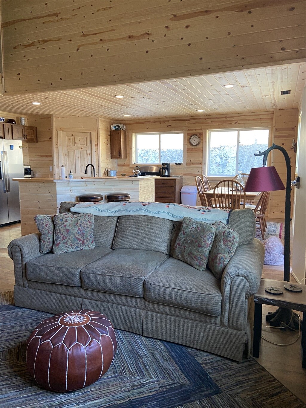 A comfortable living area features a plush sofa with decorative pillows, positioned on a patterned rug. Nearby, a dining area with a wooden table and chairs is visible, along with a practical kitchen space, illuminated by natural light from large windows.