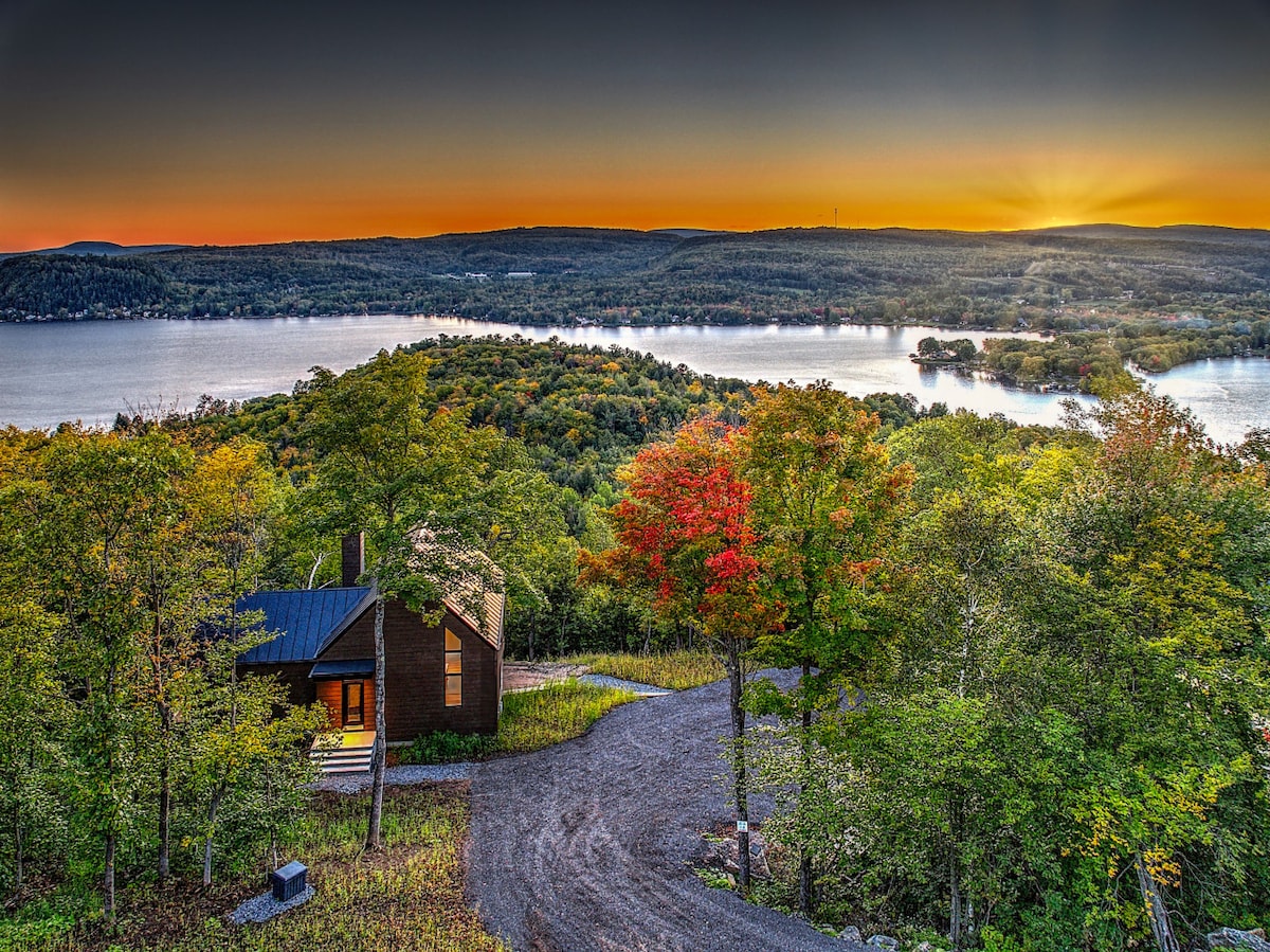 A tranquil view of a chalet located on a mountainside. The image captures vibrant autumn foliage and a winding gravel driveway, leading to the chalet. In the background, a serene lake reflects the colors of the sunset, surrounded by lush greenery and distant hills.