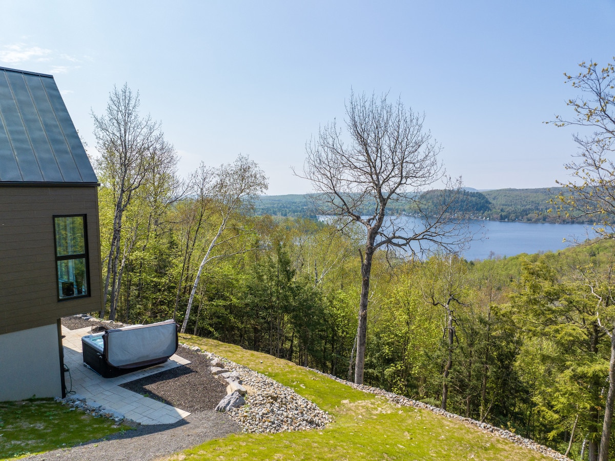 A tranquil outdoor space featuring a spa positioned on a stone patio, surrounded by lush greenery. In the background, a serene lake is visible, framed by trees and a clear blue sky, providing a peaceful retreat.