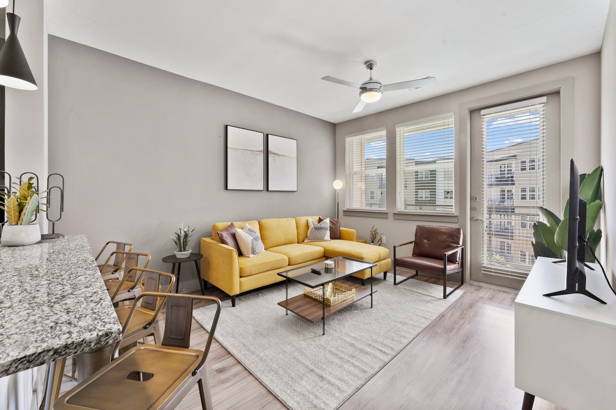 A bright living area is presented, featuring a yellow sectional sofa and a brown armchair. Large windows allow natural light to fill the space, which is accented by a light-colored area rug and a sleek coffee table. A modern fan hangs from the ceiling.