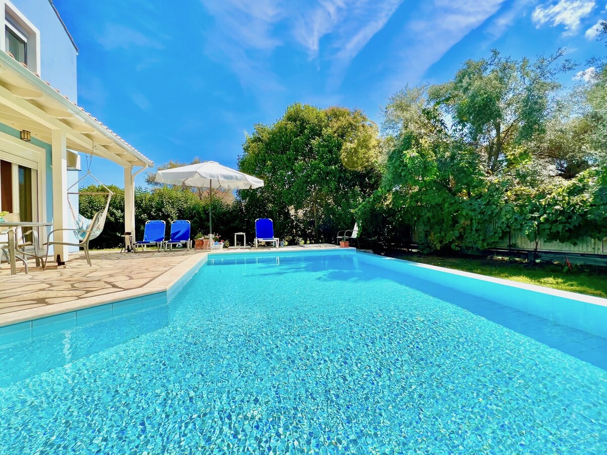 A sparkling private swimming pool reflects the clear blue sky, surrounded by lush greenery. Comfortable sunbeds and a large parasol provide shaded relaxation areas, while a patio area with seating invites outdoor dining near the water.