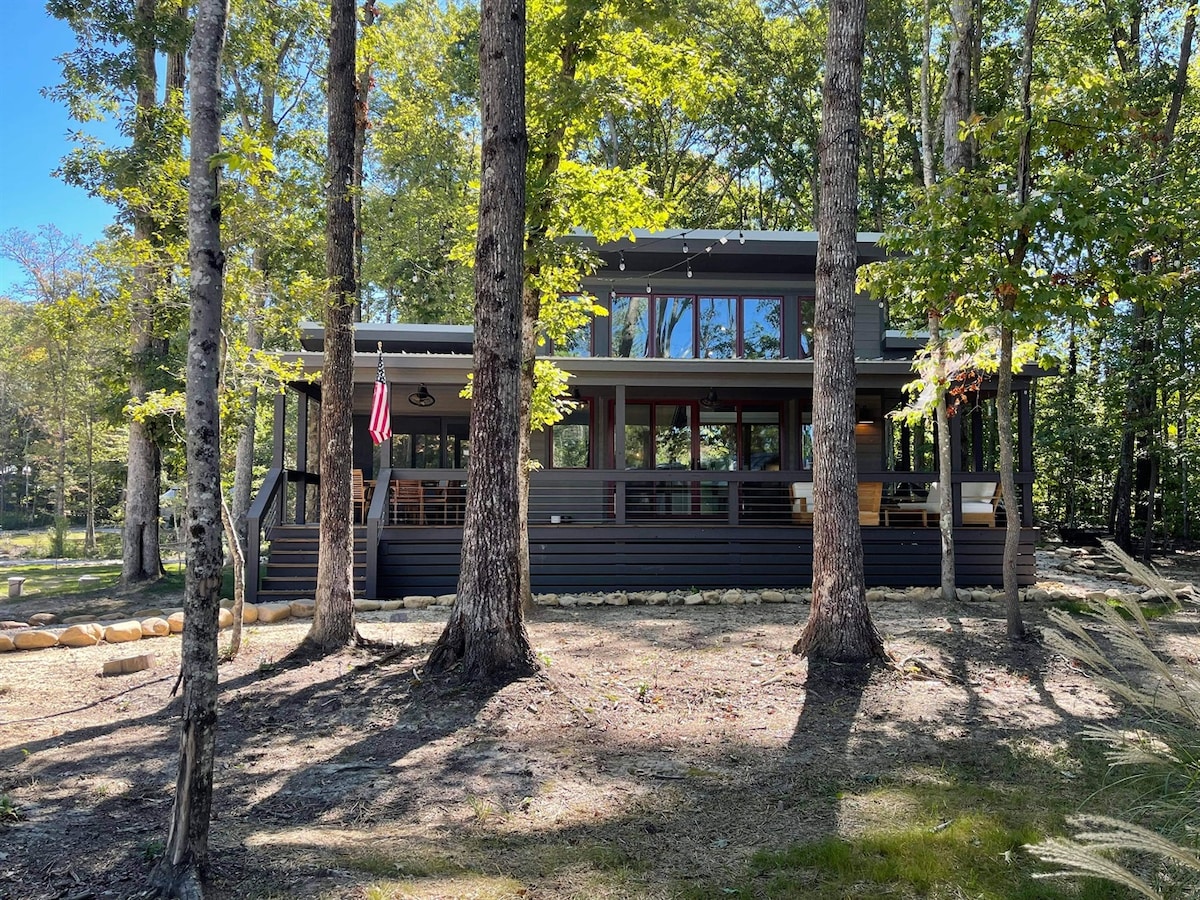 A modern home is surrounded by tall trees, showcasing a two-story design with a dark exterior. Large windows allow natural light to enter, while an American flag is displayed on the front porch. A well-maintained yard features earthy tones and stone landscaping.