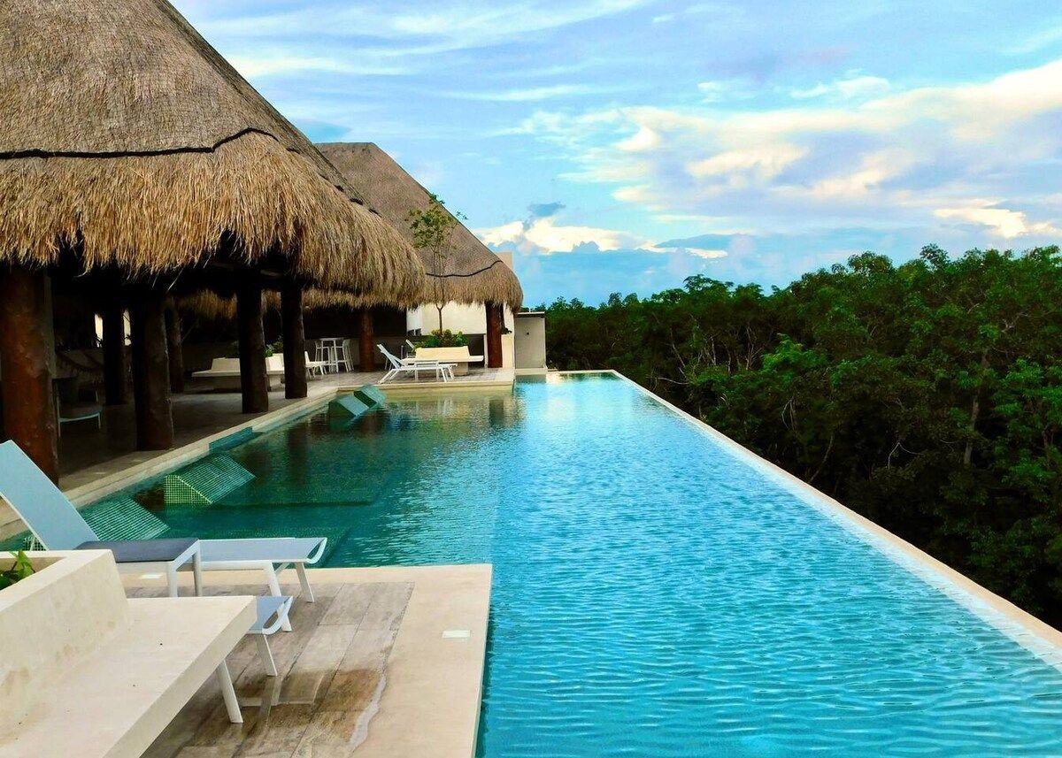 An infinity pool extends toward the horizon, bordered by thatched-roof structures. Light blue water reflects the sky, while lounge chairs are positioned on the sunlit deck, offering space for relaxation. Lush greenery is visible in the background, enhancing the natural setting.