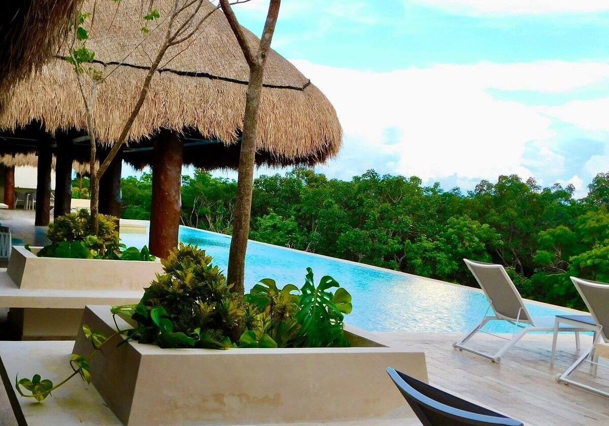 An infinity pool stretches into the lush greenery, framed by tropical plants and lounge chairs. Thatched-roof cabanas provide shaded areas for relaxation, creating a serene outdoor space. The sky reflects soft hues of blue, enhancing the connection with nature.