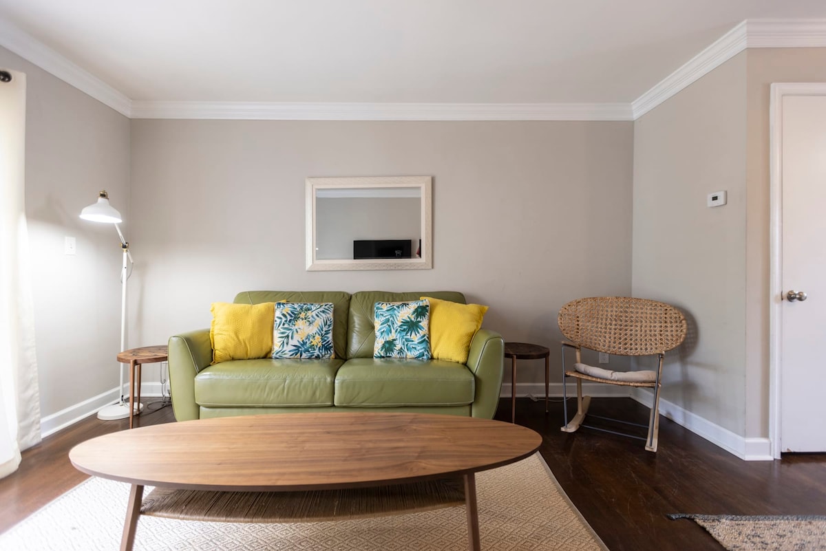 A comfortable seating area features a green sofa adorned with yellow decorative pillows. A wooden coffee table is positioned in front of the sofa, complemented by a woven chair nearby. Natural light filters through the curtains, highlighting the neutral wall tones.