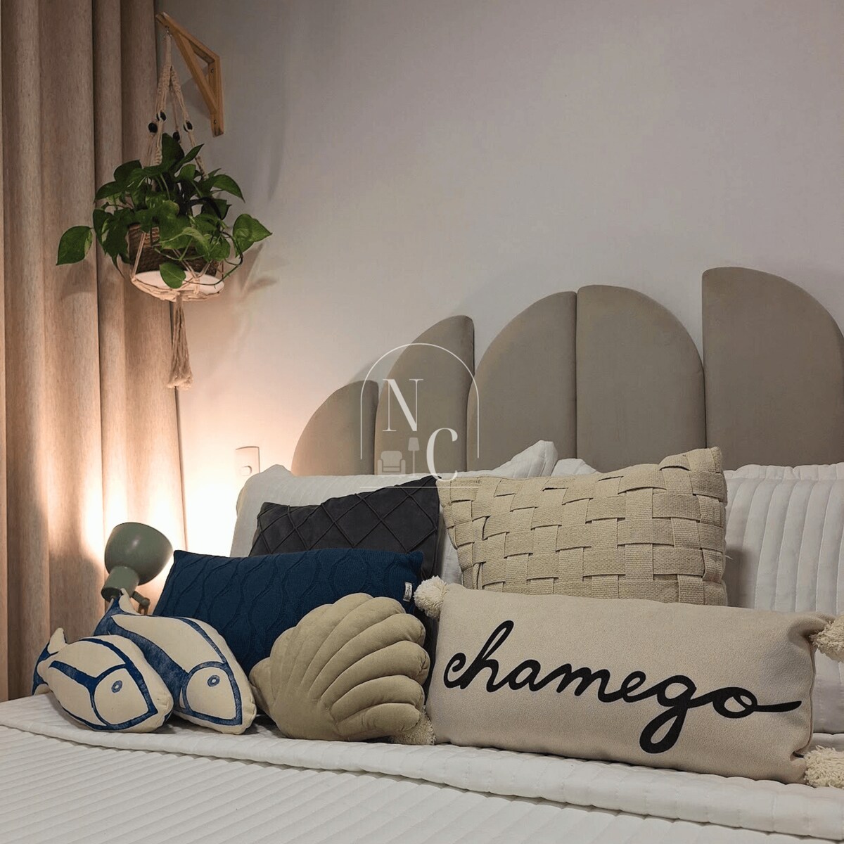 A cozy bed is decorated with various decorative pillows in soft neutral tones, including a unique pillow with the word 'chamego.' A small green plant hangs nearby, adding a touch of nature to the calming atmosphere.