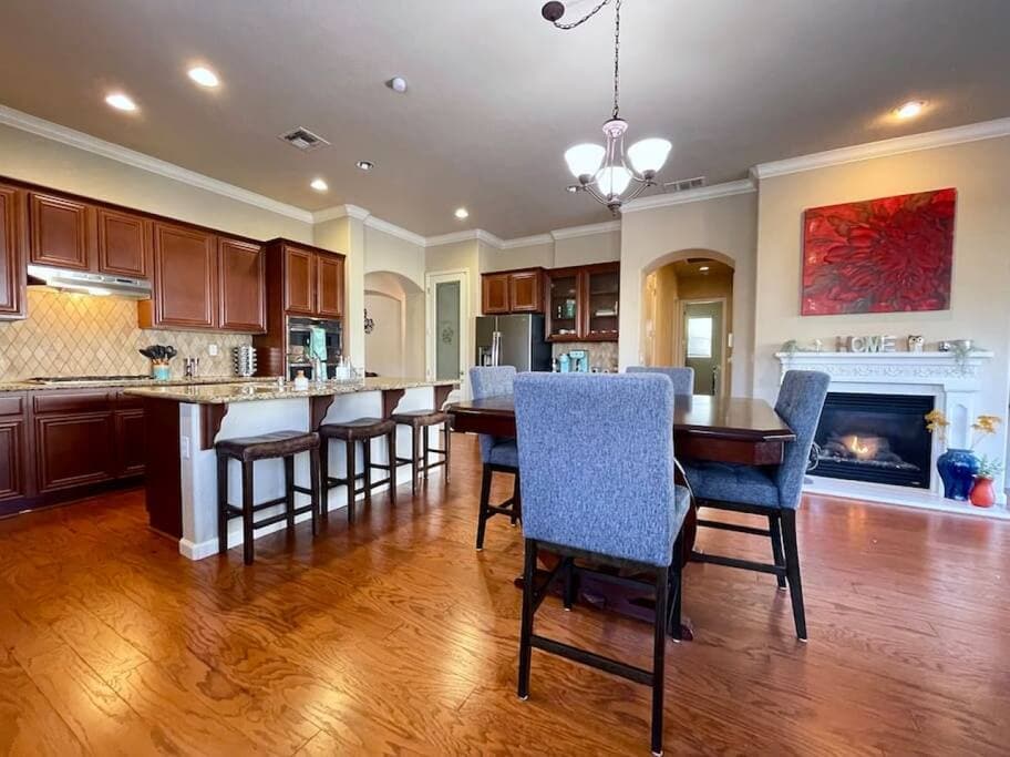 The spacious kitchen features dark wooden cabinetry and a large island with bar stools. A dining table with upholstered chairs is visible alongside a cozy fireplace. Warm lighting highlights the open layout and hardwood flooring, creating a welcoming space for gatherings.