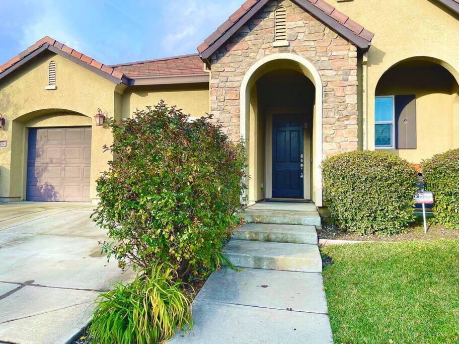 The inviting exterior of the home is showcased, featuring a stone-accented entrance and a gently curved walkway. Lush greenery borders the entrance, while the garage door blends seamlessly with the warm exterior tones. Calm skies and natural light illuminate the welcoming facade.