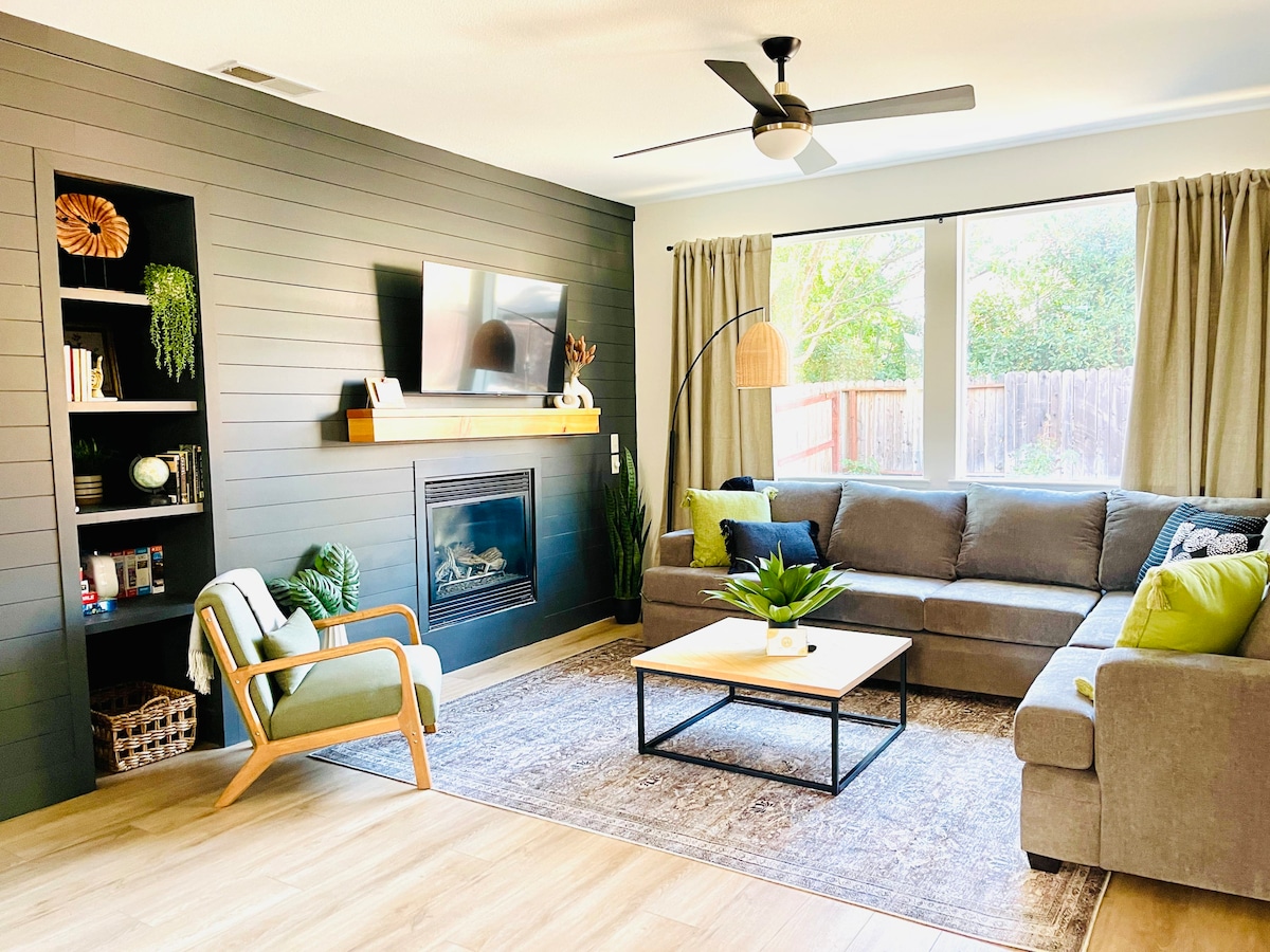 A living room is featured with a gray sectional sofa and a light wood coffee table centered on a soft area rug. A black fireplace and a television are present on the wall, complemented by a plant stand and decorative shelves. Natural light enters through large windows adorned with curtains.