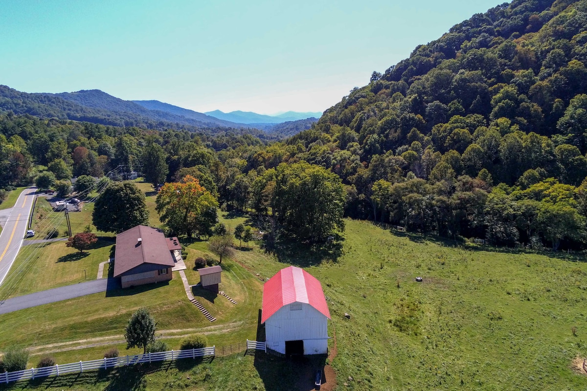 Aerial view of a spacious property surrounded by lush greenery and rolling hills. A red and white barn is positioned prominently, with a single-story house nearby, contrasting against the expansive farmland and forested mountains in the background.