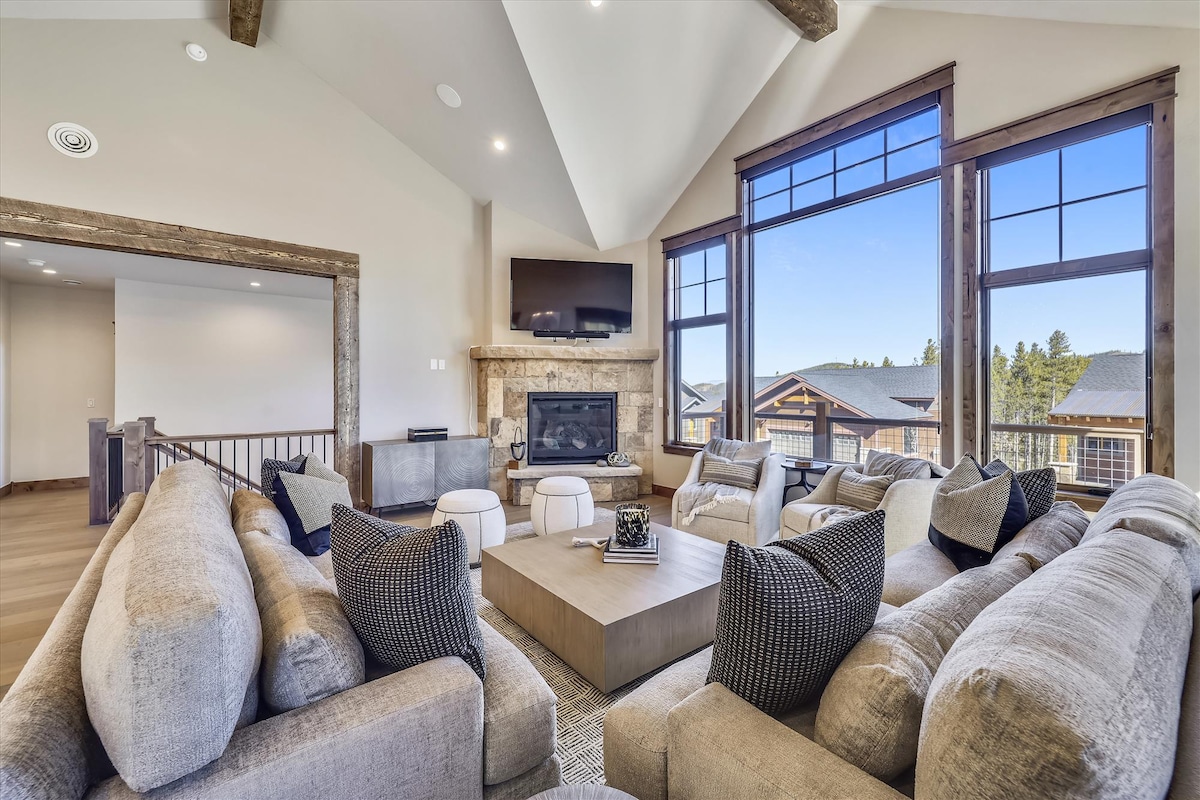 A spacious living area features a large sectional sofa and comfortable armchairs arranged around a central coffee table. Large windows provide abundant natural light and showcase mountain views. A stone fireplace and a mounted television enhance the cozy atmosphere, with wooden beams adding architectural interest.