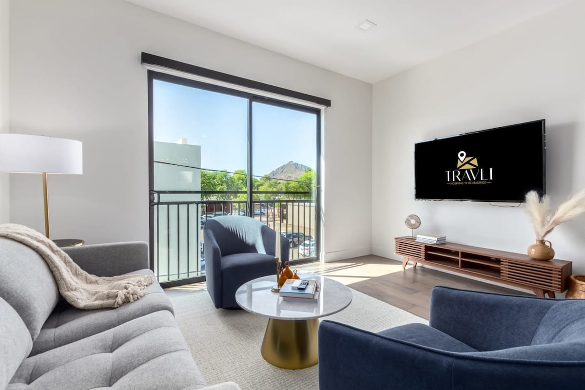 The living area features a cozy seating arrangement with a gray sofa and a plush chair, accented by a round coffee table. Large glass doors allow natural light to flood the space, offering views of the outdoors and Camelback Mountain.