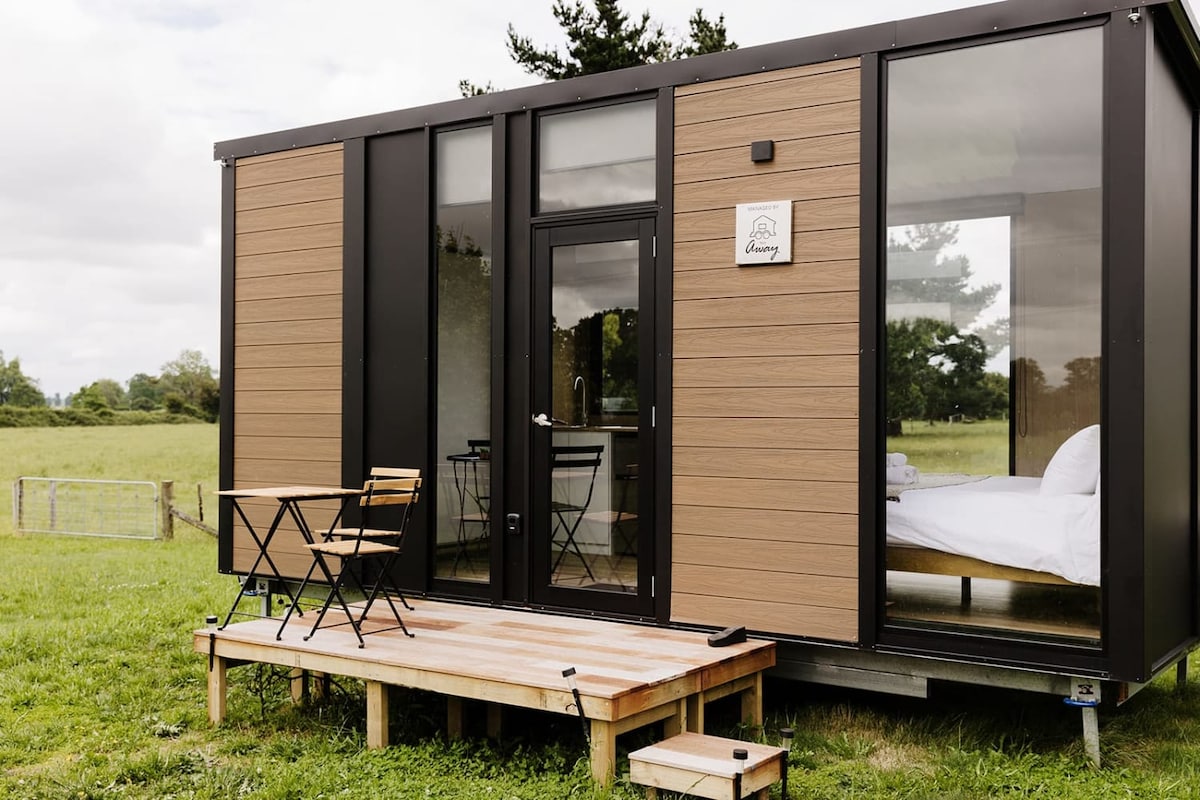 A tiny home structure showcases a modern design with large glass panels, allowing for ample natural light. The entry features a wooden deck with a small table and chairs, positioned to overlook the rural landscape, providing a serene outdoor seating area.