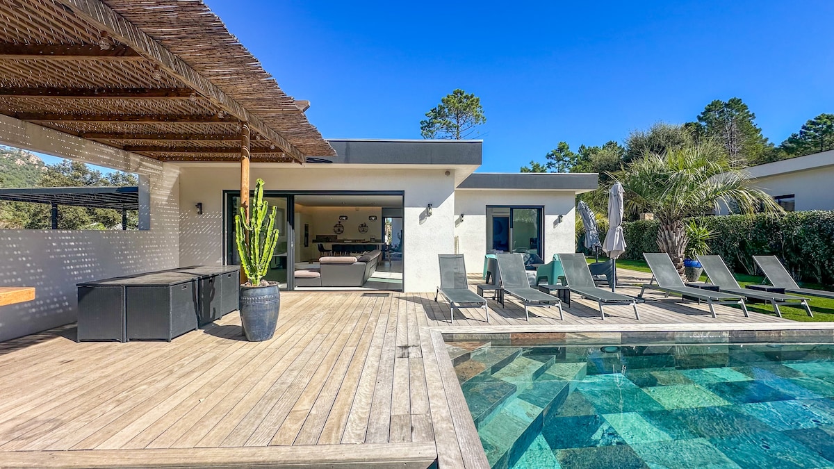 A spacious wooden terrace surrounds a private heated swimming pool, featuring sun loungers and umbrellas. The villa's glass doors provide direct access to the living area, with views of the landscaped garden and surrounding nature.