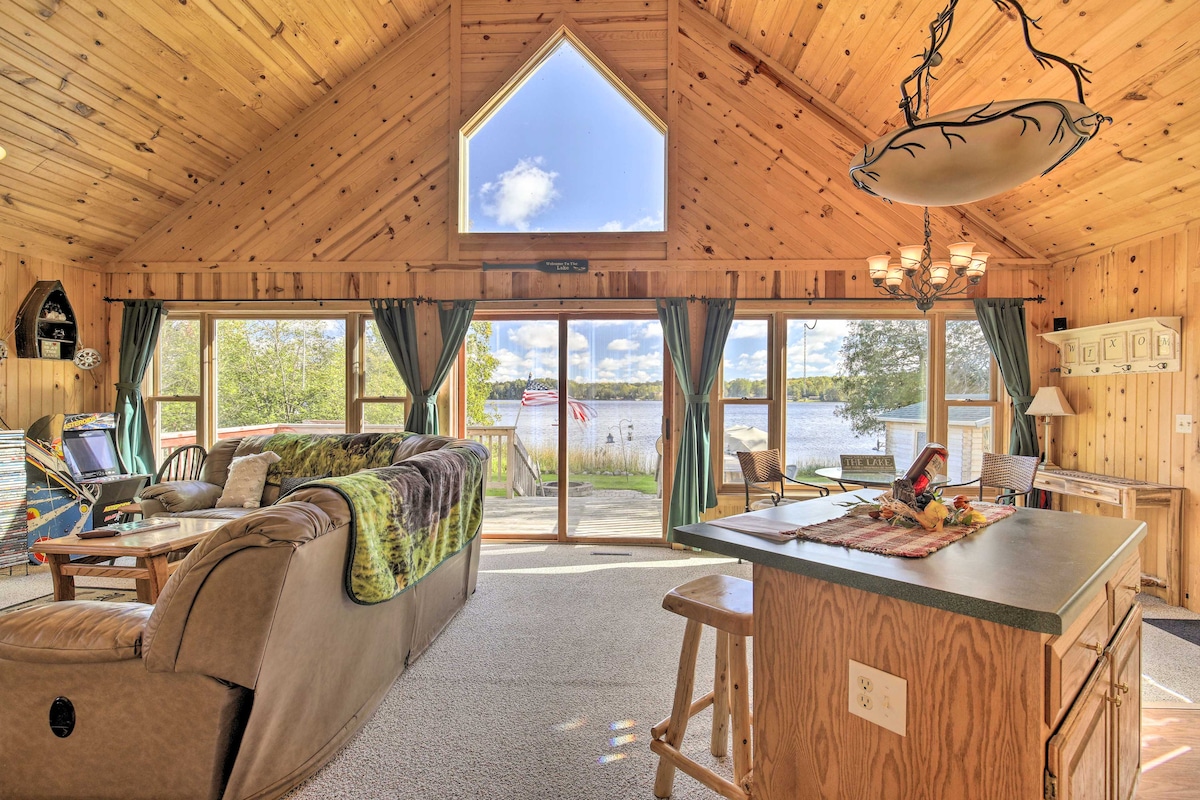An open living area features wooden walls and vaulted ceilings, creating a warm atmosphere. Large windows fill the space with natural light while providing views of the lake. Comfortably arranged seating includes a cozy sofa and a bar area with stools, inviting relaxation.