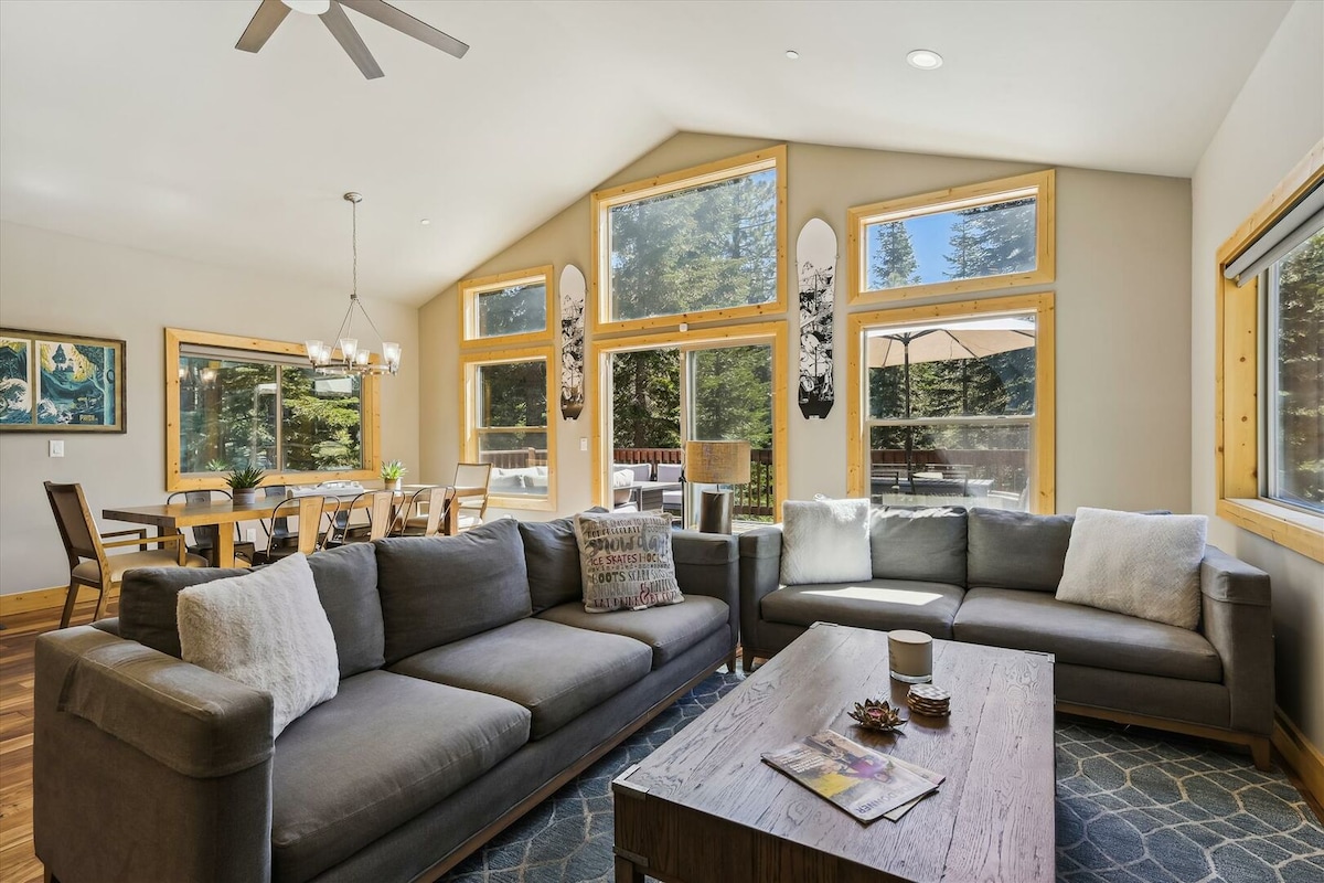 The living area features large floor-to-ceiling windows showcasing the surrounding trees, allowing natural light to fill the space. Two spacious sofas are positioned around a central wooden coffee table. A dining area with stylish seating is visible in the background, creating an inviting gathering space.