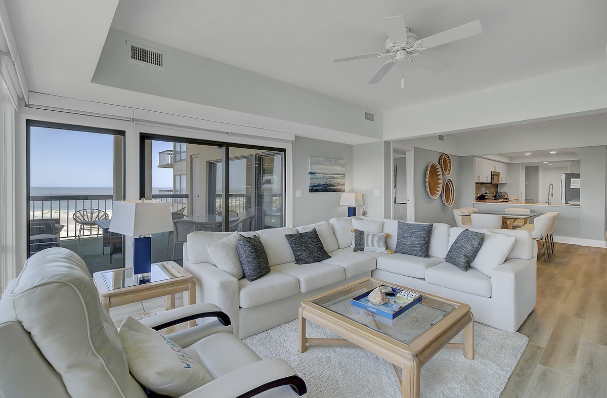 An airy living area features a spacious white sectional sofa with navy accent pillows, complemented by a coffee table set with decorative items. Large sliding glass doors provide stunning views of the ocean and access to the deck, allowing natural light to fill the room.