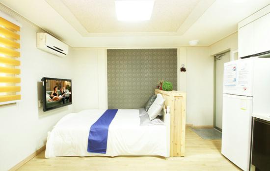 A bright and airy room features a spacious bed adorned with decorative pillows and a blue runner. A television is mounted on the wall opposite the bed. The space is complemented by light wood flooring and a modern air conditioning unit above.